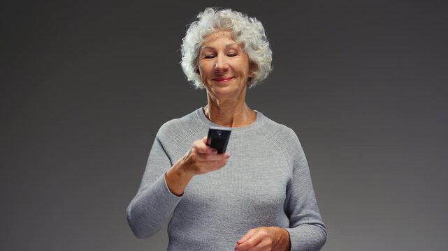 Elderly woman holding a TV remote control. Senior female switching cable channel. Old lady change broadcasting television, press button. Mature grandmother gets relax time, watching movie or news.