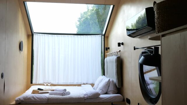 Cozy and compact bedroom inside a modern tiny house featuring light wood paneling, a comfortable bed with a breakfast tray, and a large window opening to reveal a beautiful green forest view