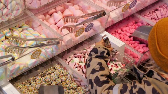 Child's hand in a leopard print jacket using tongs to select colorful sweet gummies from a pick and mix dispenser in a candy shop, choosing from a large variety of sugary treats