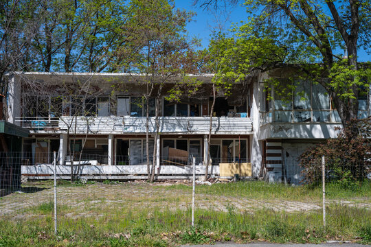 A dilapidated, two-story abandoned white concrete building with broken windows and a stripped interior stands neglected behind a fence and overgrown trees under a bright blue sky.