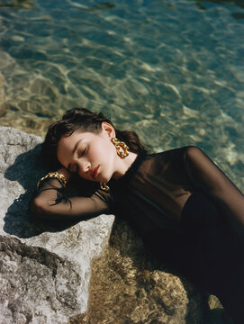 Model reclines on large stone beside clear shallow water, exuding elegance and calmness. interplay of light and water of scene