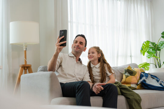 Happy father and young daughter taking a selfie with smartphone on sofa at home, Smiling dad and daughter bonding and looking at smartphone screen on grey couch in living room