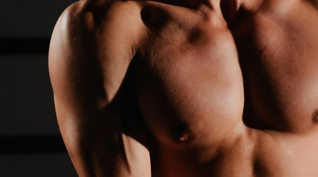 Muscular man chest and arm shown in close-up. Defined pectoral muscles and deltoid muscles visible. Physique details highlighted with dramatic lighting. Focus on powerful human form.