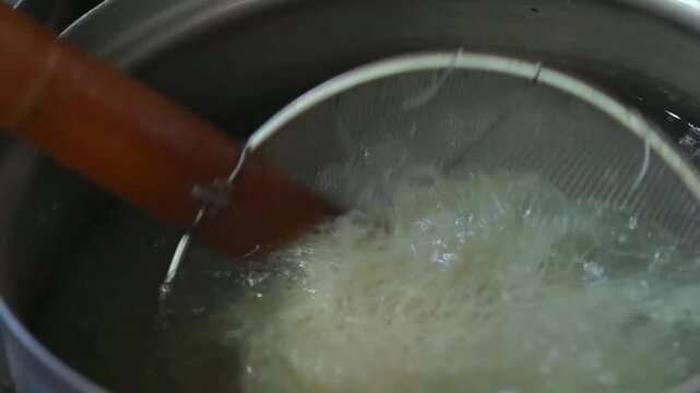 Rice vermicelli noodles boiling in hot water, close-up Asian kitchen food preparation with steam and metal strainer