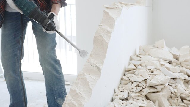Unknown male construction worker wearing red checkered shirt jeans and protective gloves, is demolishing white wall with rotary hammer drill, generating debris on the floor, close up view. Renovation