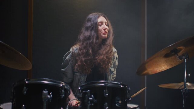 Young female drummer passionately playing a drum kit in a professional rehearsal studio, energetically striking cymbals and drums with drumsticks. Hair flying. Rock Feminist style. Woman power.