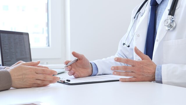 Adult aged doctor man taking notes while listening to a female patient explaining her symptoms during a medical consultation in the clinic office. Medicine and health care concept