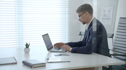 Fototapeta premium Businessman typing on laptop at office desk, analyzing financial charts and data on screen, focused on remote work productivity, planning investments and professional projects