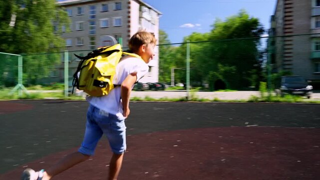 Boy runs across playground with bright yellow backpack. Girl plays nearby, chasing a ball or skipping. Sunlight glints off pavement as children enjoy outdoor time. Footage captures joyful