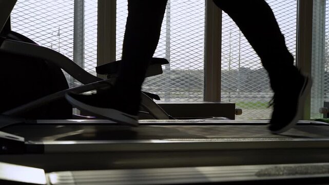 Silhouette moves steadily on a treadmill. Feet pump rhythmically, showing effort. Light filters through a mesh screen. Focus remains on steady, determined motion. Perfect for fitness or health content