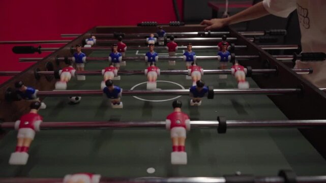 Foosball table with players in red and blue uniforms. Hands move rods to control miniature figures. Players concentrate, focused on the ball. Game moment captured with dynamic motion