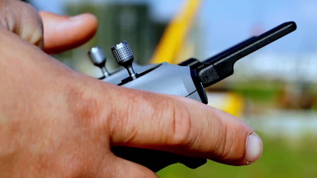 Hand grips remote control firmly. Thumb adjusts dial with precision and focus. Background hints at outdoor drone operation. Ideal for tech, drone, or outdoor hobby scenes