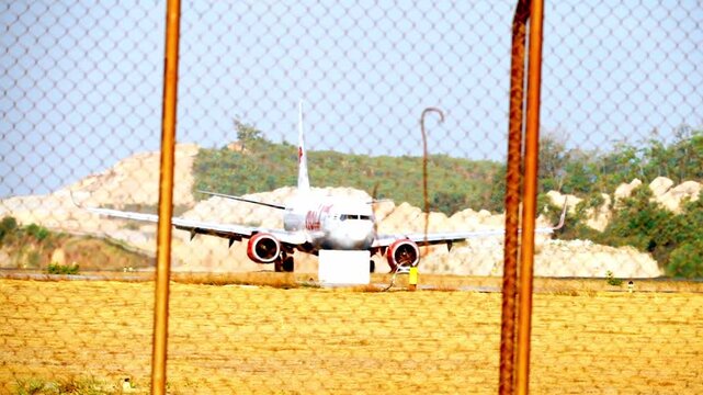 Plane sits idle behind rusted chain-link fence. Yellow grass covers ground near airfield. Mountain slope rises behind aircraft. Stillness suggests quiet, uneventful pause. Perfect for aviation