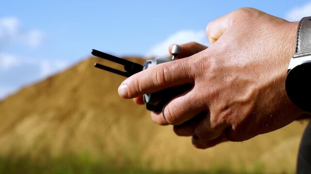 Hands grip remote, focused on drone control. Blue sky, golden earth, soft focus background. Steady hands suggest calm, precise operation. Outdoor drone piloting, likely for survey or photography