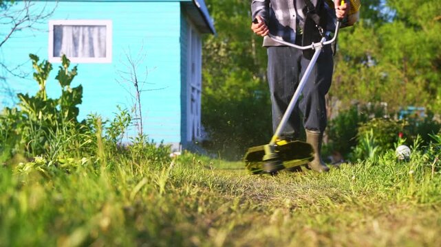 Person cuts grass with powered trimmer. Green blades slice tall weeds. Blue shed stands in background. Sunlight glints off metal handle. Motion creates soft dust clouds