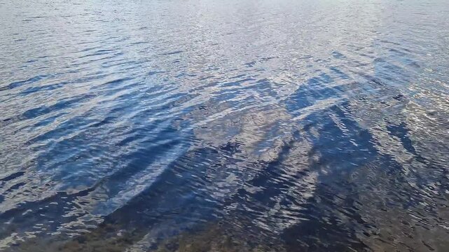 Blue water surface with light waves and reflections of the sky. Texture of water of a lake or river in daylight. The surface of the water is covered with small waves, in which the sky and clouds are r