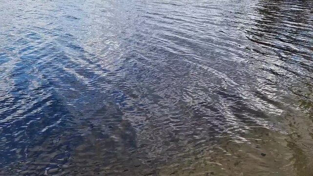 Blue water surface with light waves and reflections of the sky. Texture of water of a lake or river in daylight. The surface of the water is covered with small waves, in which the sky and clouds are r