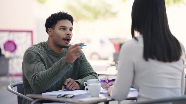 Two multiethnic university students sitting at an outdoor table studying and talking. Young man explaining a concept to his female classmate