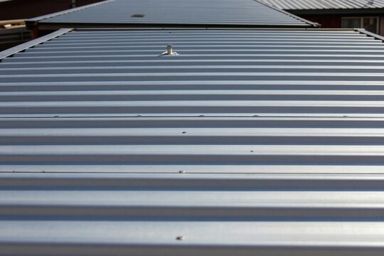 Corrugated metal roof panels, common construction material for buildings. Gray metallic surface reflects sunlight, showing clean, modern architectural detail. Material durable, often used for