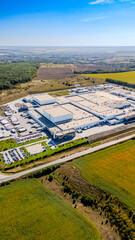 Obraz premium Vertical aerial of large logistics hub and forest landscape. High altitude bird's-eye view of a massive industrial building near green woods and yellow agricultural fields.