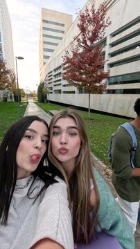 Happy young students walking together through the university campus. Two female friends are smiling and posing for a selfie video with their friends