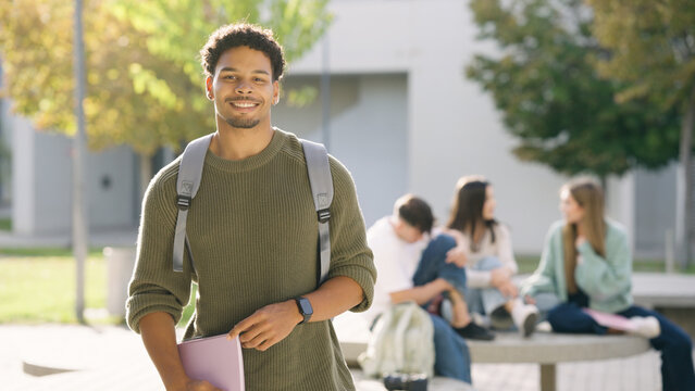 African American student smiling, holding a notebook, wearing a backpack, with classmates relaxing in the background. Studying outdoors