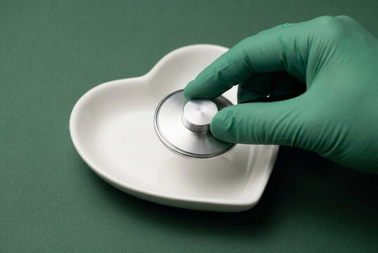 Medical hand in green glove placing silver stethoscope inside white heart shaped bowl