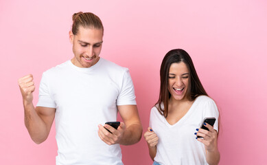 Fototapeta premium Young caucasian couple isolated on pink background using mobile phone and doing victory gesture