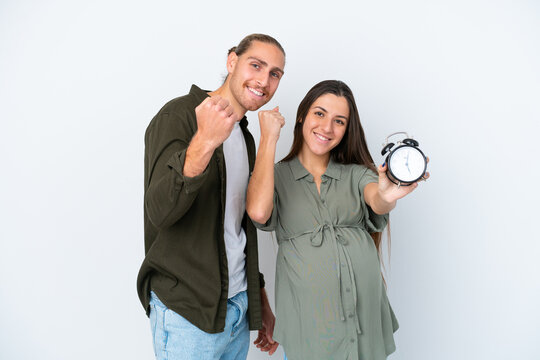 Young caucasian couple isolated on white background pregnant and holding clock while celebrate something