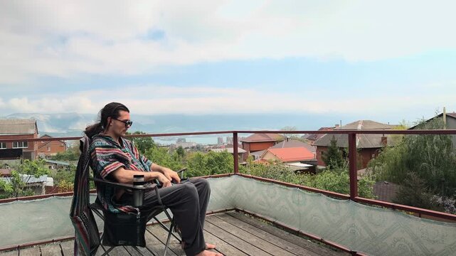 Authentic Bohemian Man with Long Hair Sitting in Folding Camping Chair on Terrace Putting on Sunglasses and Adjusting Colorful Ethnic Poncho overlooking Scenic Coastal Mountains Wide Profile Shot
