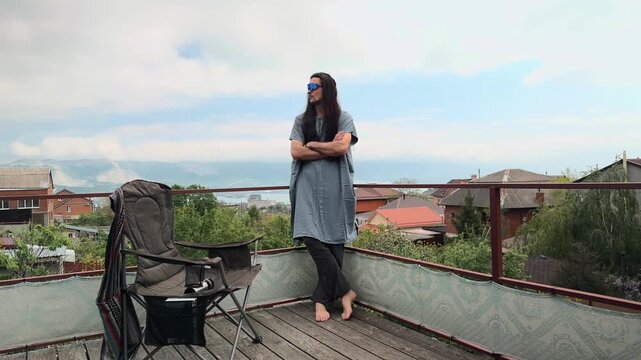 Authentic Millennial Nomad with Long Loose Hair wearing Blue Mirrored Sunglasses and Cotton Tunic Leaning on Terrace Railing Crossing Arms and Looking Aside at Coastal Village Bay Landscape Wide