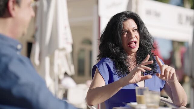 Expressive middle aged woman with long dark hair actively gesticulating while talking with a man at an outdoor cafe on a sunny day