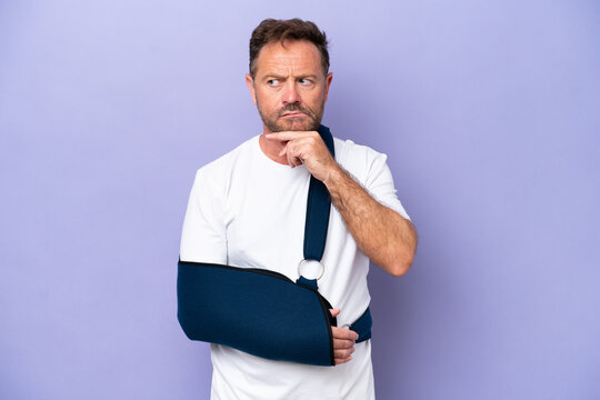 Middle age caucasian man with broken arm and wearing a sling isolated on purple background having doubts and thinking