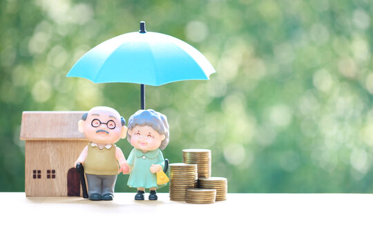 Happy elderly couple retirement life insurance and home protection concept with senior figures under blue umbrella, house model and coins on green bokeh background.