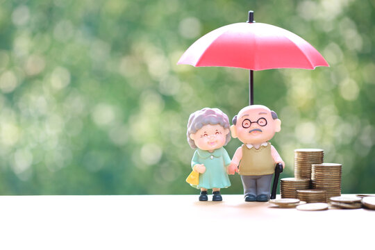 Happy senior couple retirement life insurance and financial protection concept with elderly figures under red umbrella and stacked coins on green bokeh background.