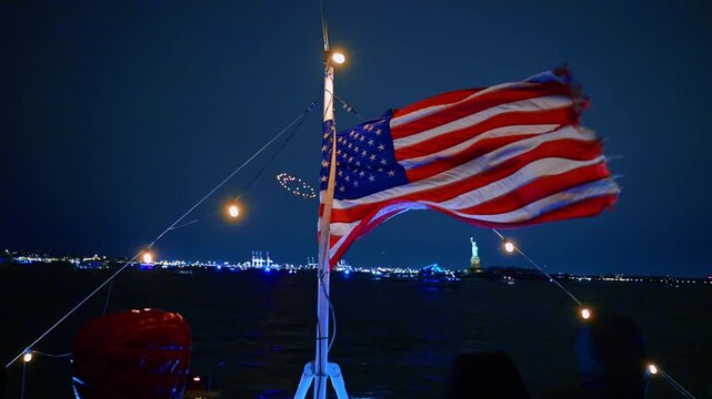 American flag flaps in the wind on the mast of the boat. People travel to the Statue of Liberty and watch the fireworks.