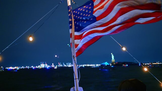 Fireworks over the docks near the Statue of Liberty. Watching the celebration of the Independence Day at night. American flag flaps in the foreground.