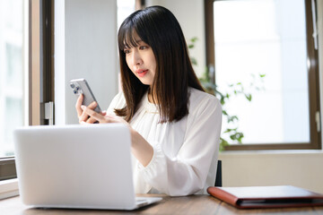 オフィスでノートパソコンとスマホを使って仕事をする日本人女性 © Yamatatsu