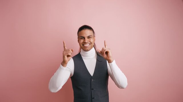 Handsome young man with long hair pointing up with both index fingers. Smiling guy indicating an empty space for advertising on a pink background