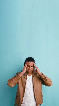 Young man with long hair feeling overwhelmed and stressed, massaging his temples due to a severe headache against a turquoise background. Vertical