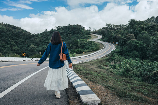 Back view of traveler asian woman with mobile phone walking on Country asphalt curved road number 3 at Nan Thailand Summer travel holiday vacation concept