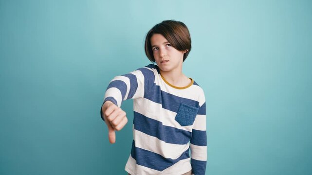 Unhappy boy in a striped shirt showing thumbs down. Displeased teen expressing disapproval and dissatisfaction on a blue background