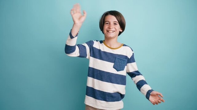 Cheerful pre teen boy in a striped shirt smiling joyfully and waving his hands in a friendly greeting against a solid blue background