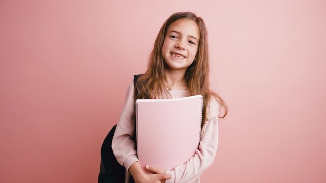 Cute little girl with a backpack holding a notebook and smiling at the camera. Studio portrait of a cheerful pupil on a pink background
