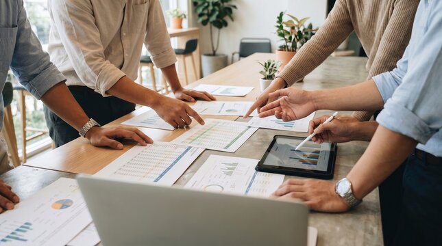 Team hands pointing at financial report and tablet showing graphs during collaborative meeting, focused planning and analysis with printed charts and laptop on wooden table
