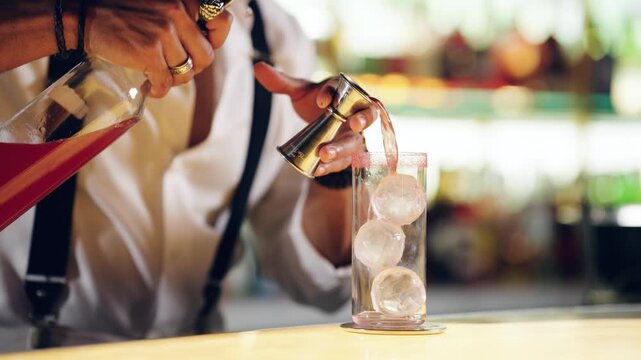 Bartender's hands preparing a refreshing red cocktail in a highball glass with spherical ice balls, using a jigger at a bar counter
