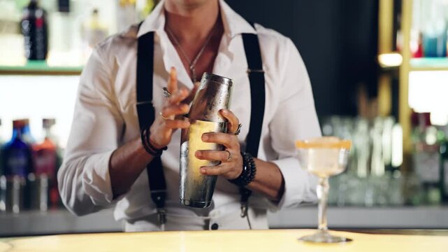 Professional bartender wearing a classic white shirt and suspenders vigorously shaking a stainless steel cocktail shaker at a stylish bar
