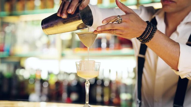Close up of a skilled bartender pouring a freshly shaken cocktail from a shaker through a strainer into a glass with a large ice cube