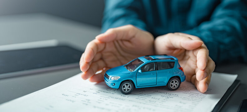 Hands protecting toy car on insurance document, symbolizing auto policy, claim coverage and financial security. Ideal for risk management, accident protection, premium plans.