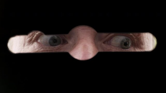 Secretive hidden man darts his eyes around while looking through keyhole of lever lock on black door close-up